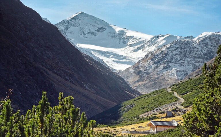 Fot vom Weitwanderweg E5 mit einem verschneiten Gipfel und einer Hütte im Grünen.